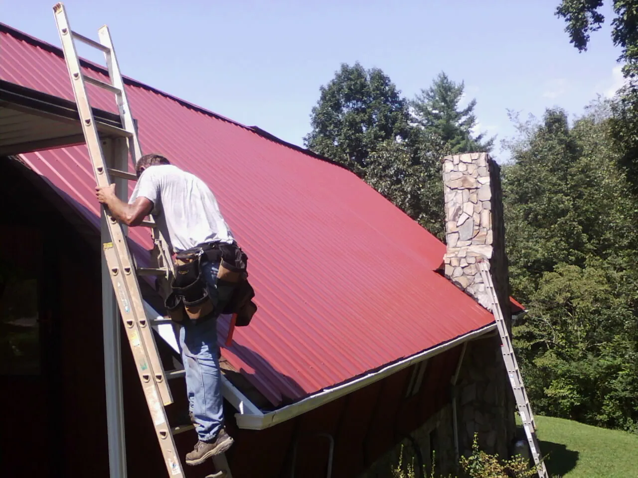 Roofer on a ladder beside a red metal roof and stone chimney — Soffit & Fascia Repair by Roofing Brothers in Garwin
